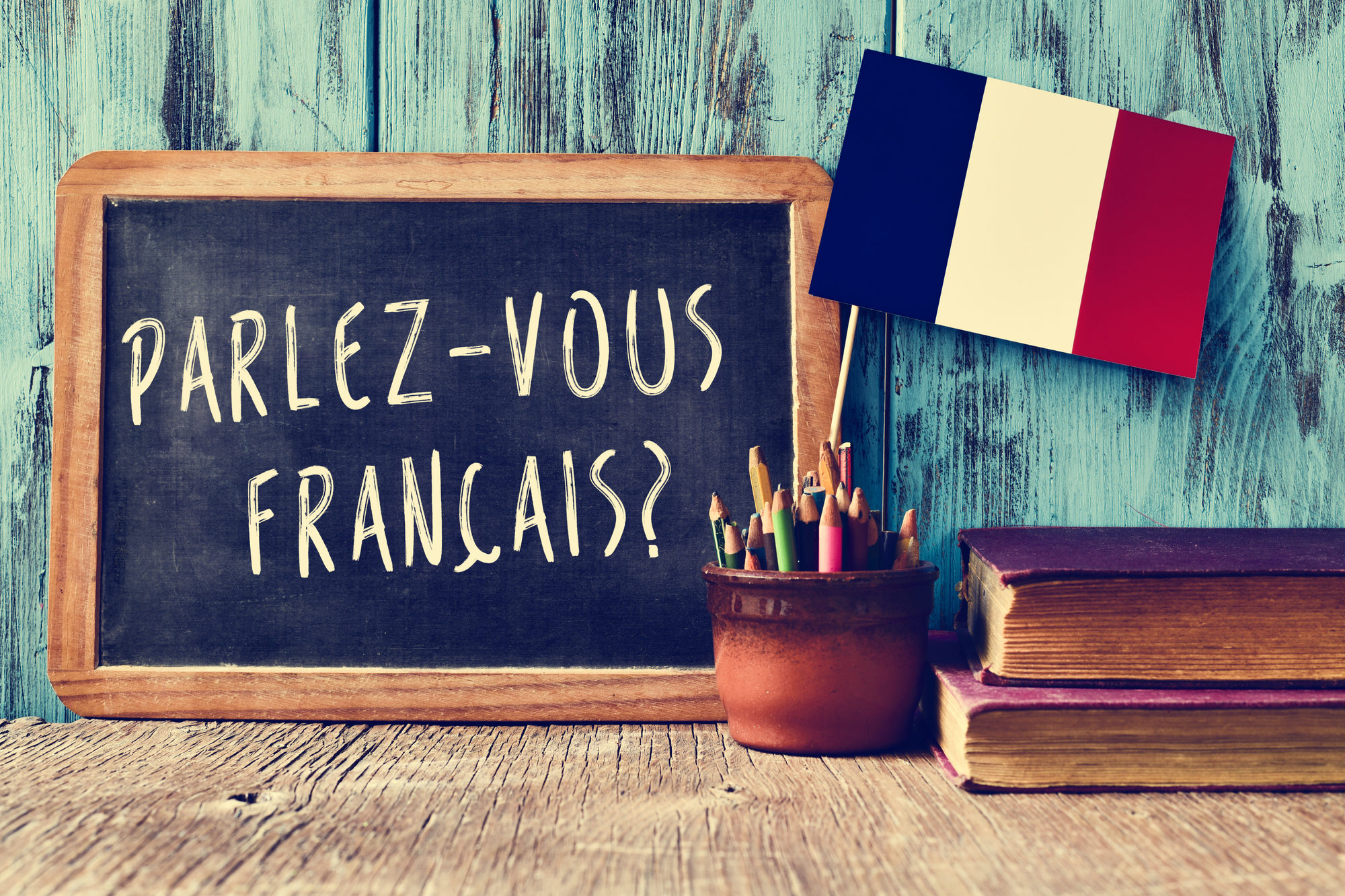 a chalkboard with the question parlez-vous francais? do you speak french? written in french, a pot with pencils and the flag of France, on a wooden desk, Growing Up French Banner